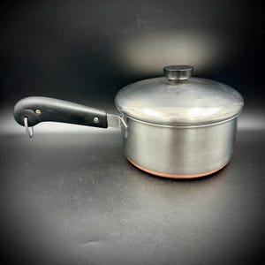 May include: A silver-toned metal saucepan with a copper-colored base, featuring a black handle with a metal hanging loop and a matching silver-toned lid with a black knob. The pot has a cylindrical body and a sturdy, riveted handle attachment.