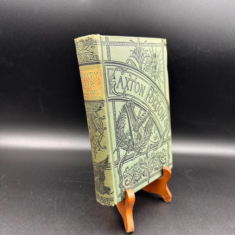 May include: A vintage book titled "Vanity Fair" by Thackeray, Caxton Edition, displayed on a wooden stand. The book has a light green cover with black decorative illustrations and text. The spine is tan with gold lettering.