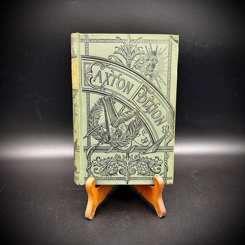 May include: A vintage book with a light green cover and the title "Caxton Edition" in a decorative font. The cover features ornate black illustrations of a bird, foliage, and other designs. The book is displayed on a small wooden stand.