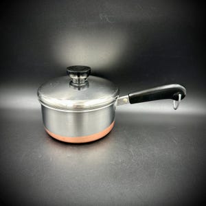 May include: A stainless steel saucepan with a copper-colored base, featuring a matching stainless steel lid with a black knob. The pan has a long black handle with a metal hanging loop attached to the end, providing a functional design for cooking.