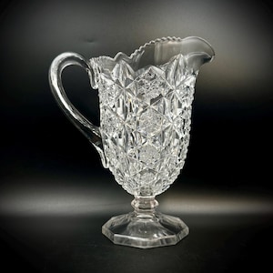 May include: Clear glass pitcher with a decorative, textured pattern. The pitcher features a handle and a scalloped rim. The base is a hexagonal shape. The pitcher is likely used for serving beverages.