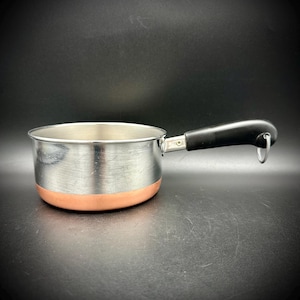 May include: A small saucepan with a polished stainless steel interior and exterior walls, featuring a copper-colored base. The pan has a black handle attached with a silver metal rivet and includes a small metal loop at the end of the handle.