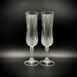 May include: A pair of clear crystal champagne flutes featuring a tall, slender bowl with vertical fluting at the top and a detailed diamond-cut pattern at the base. Each glass has a multi-faceted stem and a circular foot for stability.