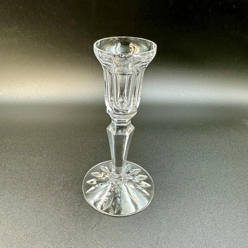 May include: A clear crystal candlestick holder with a flared top and a faceted stem. The base is round with a starburst design. The candlestick is designed to hold a single candle.