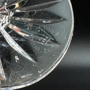 May include: Close-up of a clear crystal glass with a starburst design. The glass has a textured surface and the words "AYTON ENG" are etched into the glass. The glass is on a black background.