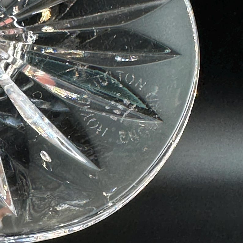 May include: Close-up of a clear crystal glass with a starburst design. The glass has a textured surface and the words "AYTON ENG" are etched into the glass. The glass is on a black background.