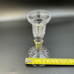 May include: A clear, faceted crystal candlestick holder with a round base and a flared top. The base has a starburst design. The candlestick is approximately 6 cm tall, as measured by a ruler.