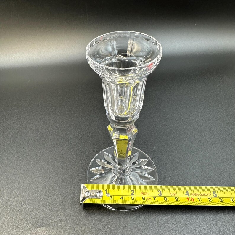 May include: A clear, faceted crystal candlestick holder with a round base and a flared top. The base has a starburst design. The candlestick is approximately 6 cm tall, as measured by a ruler.