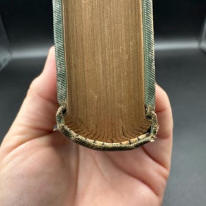 May include: Close-up of a vintage book's spine and pages. The pages are aged with a golden-brown hue, while the spine is covered in a textured, green fabric. The book's edges show wear, suggesting it's an antique.
