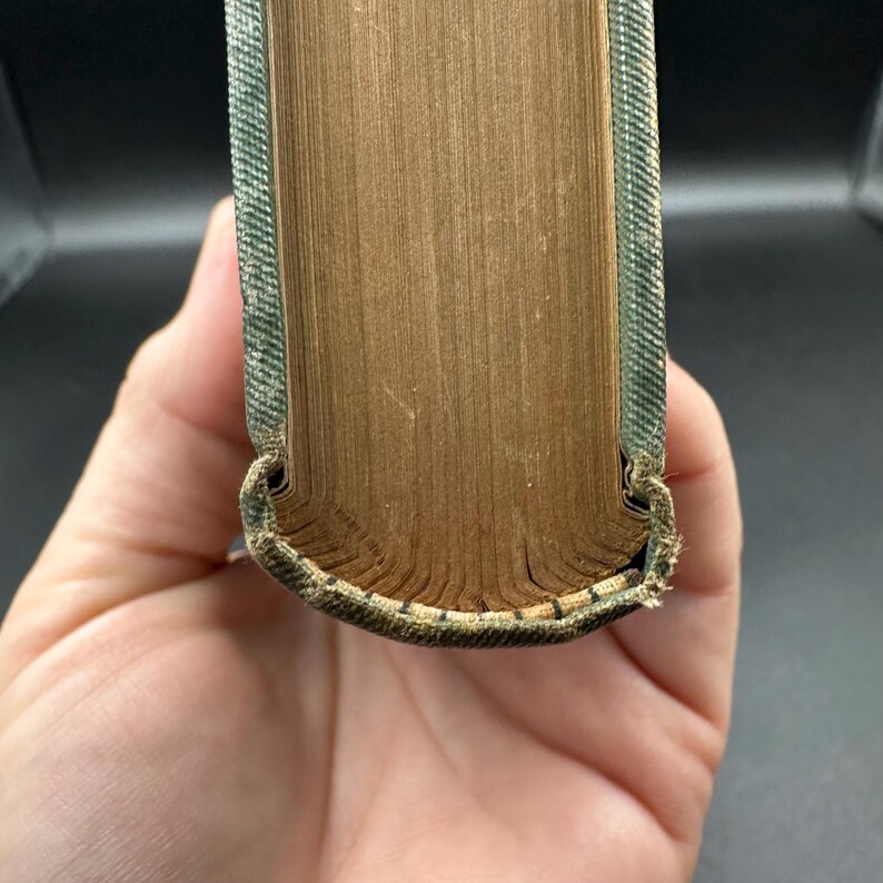 May include: Close-up of a vintage book's spine and pages. The pages are aged with a golden-brown hue, while the spine is covered in a textured, green fabric. The book's edges show wear, suggesting it's an antique.