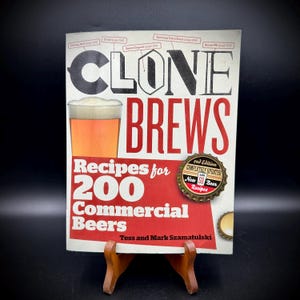 May include: A book titled "Clone Brews" with a glass of beer on the cover. The cover is white and red with the text "Recipes for 200 Commercial Beers". A bottle cap with text is on the cover. The book is on a wooden stand.