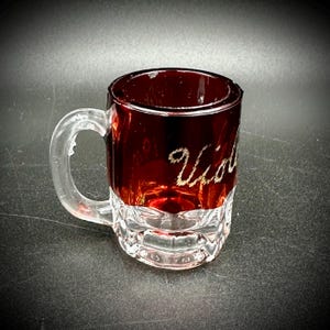 May include: A small, clear glass mug with a handle. The top portion is a deep red color, with the word "Violet" in gold script. The bottom half of the mug is clear with a textured design.