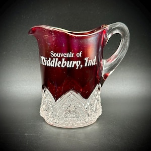 May include: A small, ruby-red glass pitcher with a clear handle and a decorative, cut-glass base. The pitcher is inscribed with the words "Souvenir of Middlebury, Ind." in white lettering. The pitcher is likely a vintage souvenir item.