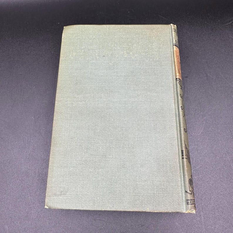 May include: A vintage book with a light green cover. The spine is decorated with ornate designs and a strip of tan paper. The book is closed and angled, showing the cover and spine against a dark background.