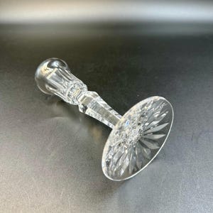 May include: A clear crystal candlestick holder with a round base and a faceted stem. The base features a starburst design. The candlestick holder is lying on a dark surface.