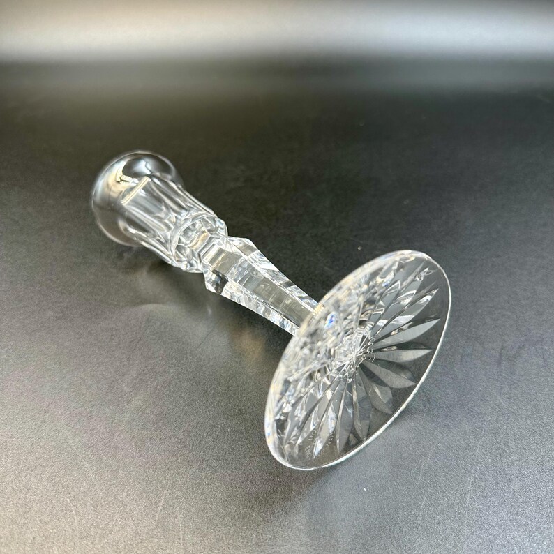 May include: A clear crystal candlestick holder with a round base and a faceted stem. The base features a starburst design. The candlestick holder is lying on a dark surface.