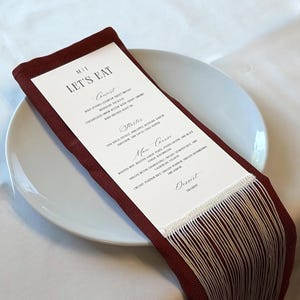 Peut inclure: Une carte de menu rectangulaire intitulée LET'S EAT avec du texte noir sur fond blanc, placée sur une serviette en tissu rouge foncé avec de longues franges claires en bas, reposant sur une assiette blanche. Le menu liste les plats Couvert, Starter, Main Course et Dessert.