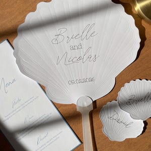 May include: White seashell-shaped fan with the names "Brielle and Nicolas" and the date "09.02.2026" printed on it. A menu and place cards with names are also visible. The fan has a plastic handle.
