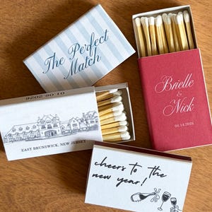 Könnte beinhalten: Mehrere Streichholzschachteln mit individuellen Designs. Eine Schachtel trägt den Schriftzug "The Perfect Match" auf einem blau-weiß gestreiften Hintergrund. Eine andere ist rot mit "Brielle & Nick" und dem Datum 04.14.2026. Eine dritte Schachtel zeigt eine Gebäudeillustration und den Text "East Brunswick, New Jersey". Eine vierte Schachtel trägt den Text "Cheers to the new year!"