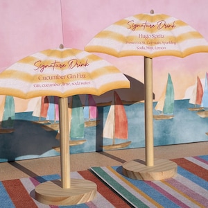 K&ouml;nnte beinhalten: Zwei Miniatur-Strandschirme aus Holz als Getr&auml;nkekarten. Der linke Schirm zeigt Signature Drink, Cucumber Gin Fizz, Gin, cucumber, lime, soda water. Der rechte zeigt Signature Drink, Hugo Spritz, Prosecco, St. Germain, Sparkling Soda, Mint, Lemon.