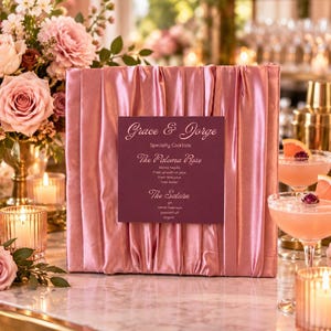 May include: A square menu sign featuring a dark maroon card with white text reading Grace & Jorge Specialty Cocktails, The Paloma Rose, and The Saturn, mounted on a pleated, shimmering dusty pink satin fabric backing.