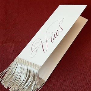 May include: A white folded card featuring the word Vows written in elegant dark script lettering. The bottom edge of the card is adorned with a long, light-colored fringe made of thin, straight threads that hang down from the paper surface.