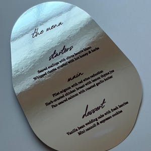 May include: A teardrop-shaped menu card with a reflective silver metallic finish. The text is printed in black, listing sections for starters, main, and dessert, including items like seared scallops, filet mignon, and vanilla bean wedding cake.