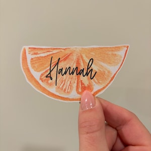 Puede incluir: Una pegatina con forma de gajo de naranja, con el nombre "Hannah" escrito en cursiva. La pegatina tiene un efecto acuarela con tonos naranja y blanco. La pegatina está sujeta por una persona.