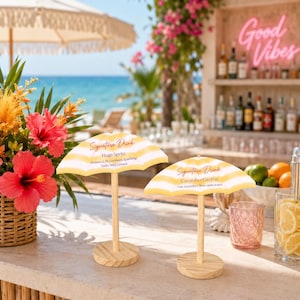 K&ouml;nnte beinhalten: Zwei Miniatur-Cocktailschirm-Schilder mit gelb-wei&szlig;en Streifen auf Holzst&auml;ndern. Das linke Schild tr&auml;gt die Aufschrift Signature Drink Hugo Spritz Prosecco St. Germain Sparkling Soda Mint Lemon. Das rechte Schild tr&auml;gt die Aufschrift Signature Drink Cucumber Gin Fizz Gin cucumber lime soda water.