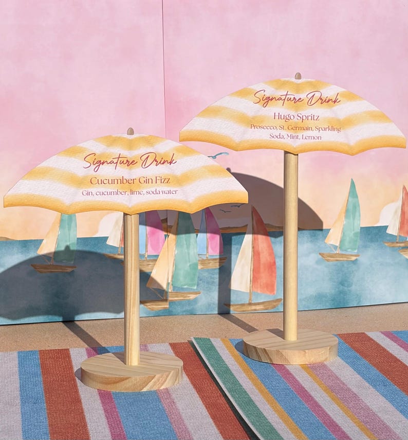K&ouml;nnte beinhalten: Zwei Miniatur-Strandschirme mit gelb-wei&szlig;en Streifen, jeweils mit einem Holzstab und einem Holzsockel. Der linke Schirm zeigt den Text Signature Drink Cucumber Gin Fizz Gin, cucumber, lime, soda water. Der rechte zeigt Signature Drink Hugo Spritz Prosecco, St. Germain, Sparkling Soda, Mint, Lemon.