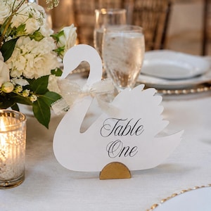Puede incluir: Un letrero de mesa con forma de cisne blanco que presenta el texto negro Table One impreso en su lateral. El letrero está adornado con una pequeña cinta de tela de color claro atada alrededor del cuello del cisne y una base dorada semicircular.