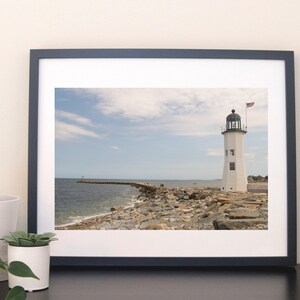 May include: A framed photograph of a white lighthouse with a green roof and a small American flag on a rocky shore. The lighthouse is on a small island connected to the mainland by a rocky path. The sky is blue with white clouds.
