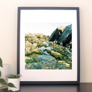 Op de afbeelding: Ingelijste foto van een rotsachtige kustlijn met helder water en groene algen. De afbeelding staat in een zwart frame en wordt weergegeven op een neutraal gekleurde muur. De scène legt een natuurlijk kustlandschap vast met grote rotsen en een kalme zee.