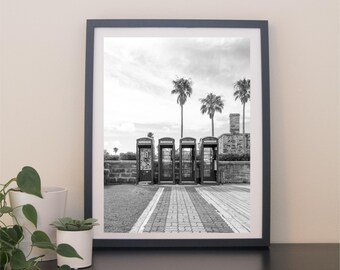 May include: Black and white framed print featuring a row of vintage telephone booths. Palm trees and a cloudy sky are visible in the background. The booths are aligned on a brick and wooden pathway.