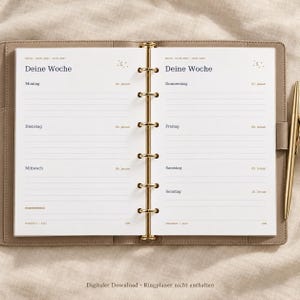 Op de afbeelding: Een open ringbandplanner met een beige kaft en goudkleurige ringen. De pagina's bevatten een weekindeling in het Duits met de tekst "Deine Woche" en "Wochenfokus", inclusief de data van 4 tot 10 januari 2027. Een gouden pen ligt aan de rechterkant.