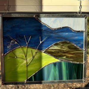 May include: A stained glass window depicting a landscape with a green hill, a brown hill, and a blue sky. A small tree with thin branches is in the foreground. The window is framed in silver metal.