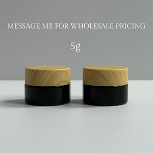 May include: Two small, black glass jars with light wood-grain lids. The jars are labeled with "5g" in a simple font. The text "MESSAGE ME FOR WHOLESALE PRICING" is above the jars.