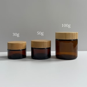 May include: Three amber glass jars with wooden lids. The jars are labeled 30g, 50g, and 100g. The jars are cylindrical and have a minimalist design, suitable for storing cosmetics or other small items.