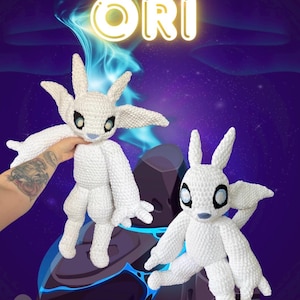 May include: Two white crochet Ori plushies with large ears, wings, and black eyes are displayed against a purple and blue background. The word "ORI" is written in large, glowing letters at the top. The text "BY : GEEKMOTHER" is at the bottom.