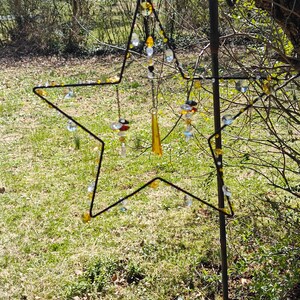 Peut inclure: Un carillon en forme d'étoile en métal avec des perles de verre transparentes et ambrées qui pendent. Le carillon est accroché à un crochet en métal noir et est suspendu à un poteau en métal noir.