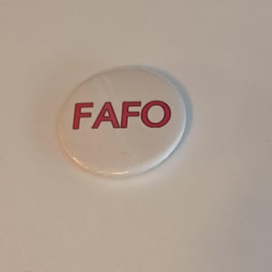 Puede incluir: Un botón blanco con la palabra "FAFO" impresa en rojo.