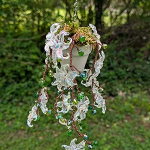 May include: A hanging white ceramic planter filled with moss, decorated with clear, iridescent flower-shaped beads and green crystal accents. The beads are strung on copper wire, creating a cascading floral design. The planter hangs from a silver chain.
