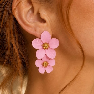 May include: A pair of drop earrings featuring two stacked light pink flower petals with textured surfaces and a small, detailed gold-toned center. The floral design consists of a larger five-petaled flower positioned above a smaller five-petaled flower.