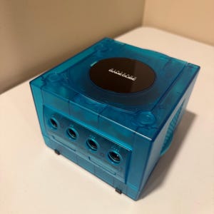 May include: A transparent blue Nintendo GameCube console. The console has a circular disc tray with the words "NINTENDO GAMECUBE" printed on it. The console has four controller ports and a handle on the side.
