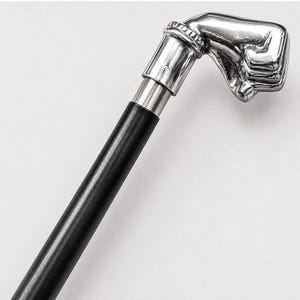 Op de afbeelding: Een wandelstok met een gepolijst zilverkleurig handvat in de vorm van een gebalde vuist. Het handvat is bevestigd aan een gladde, cilindrische zwarte schacht met een metalen kraag die de twee delen van de stok met elkaar verbindt.