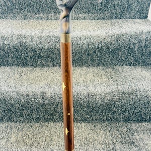Op de afbeelding: Een houten wandelstok met een decoratieve kattenkop handvat. Het handvat is zilver en goud, en de schacht is bruin met gouden accenten. De stok is ongeveer 91 cm lang.