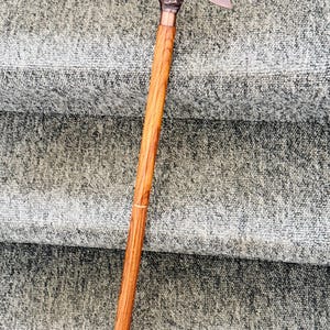 Puede incluir: Bastón de madera con un mango en forma de cabeza de conejo color cobre. El bastón tiene un eje largo y delgado de color marrón claro con un acento de cobre cerca de la parte superior. El mango está detallado con los rasgos del conejo y una oreja curvada.