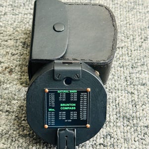 Peut inclure: Une boussole Brunton noire avec un étui en cuir. La boussole a un cadran circulaire avec des graduations et un tableau des sinus naturels. L'étui est noir et rectangulaire avec une fermeture à bouton-pression.
