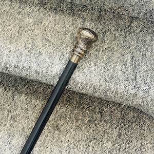 May include: A black walking cane with a decorative, ornate brass handle. The handle has a detailed floral design. The cane is made of wood and has a brass collar. The cane is approximately 36 inches long.