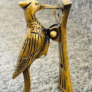 May include: A bronze-coloured metal woodpecker ornament, intricately detailed with feather markings and a long beak, perched on a tree branch. This decorative sculpture is likely for home decor.